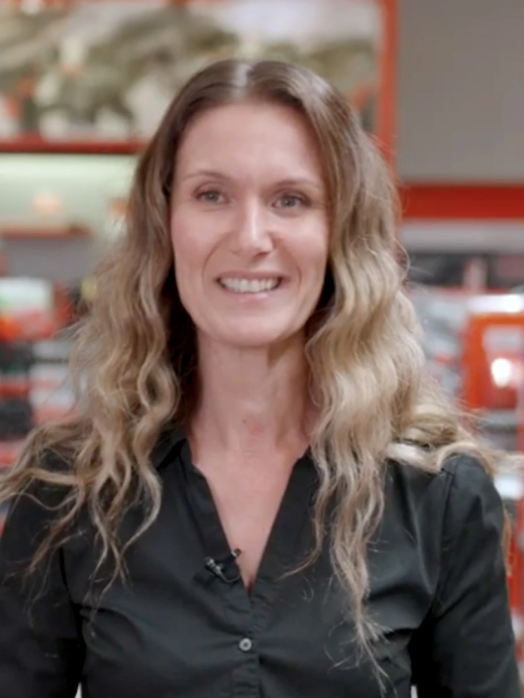 Femme souriante aux cheveux bouclés longs portant une chemise noire, avec un fond intérieur flou rouge et gris.
