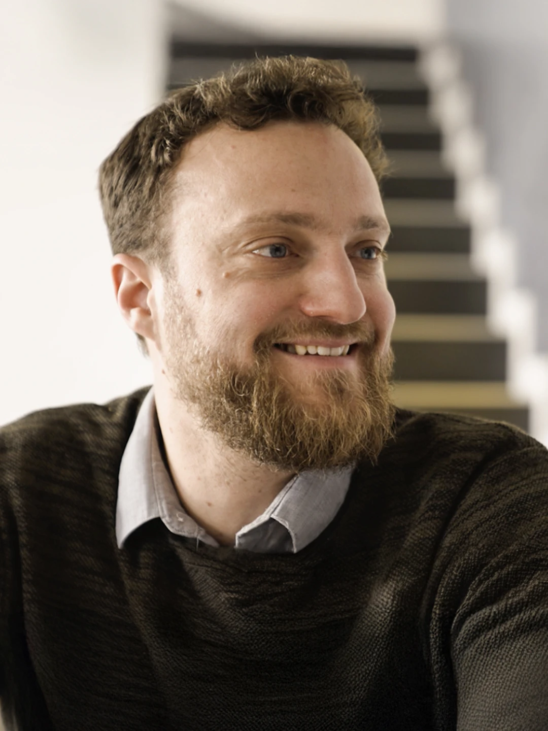 Homme souriant portant un pull sombre avec une chemise grise, en arrière-plan un escalier flou.