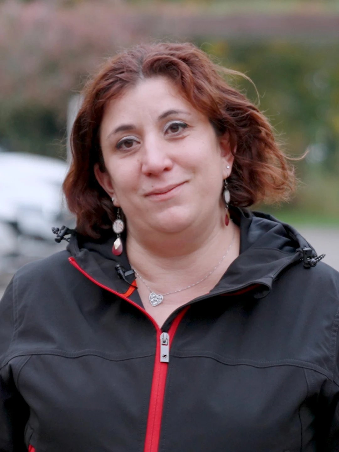 Femme aux cheveux roux courts portant une veste noire avec une fermeture éclair rouge, souriant légèrement en extérieur.