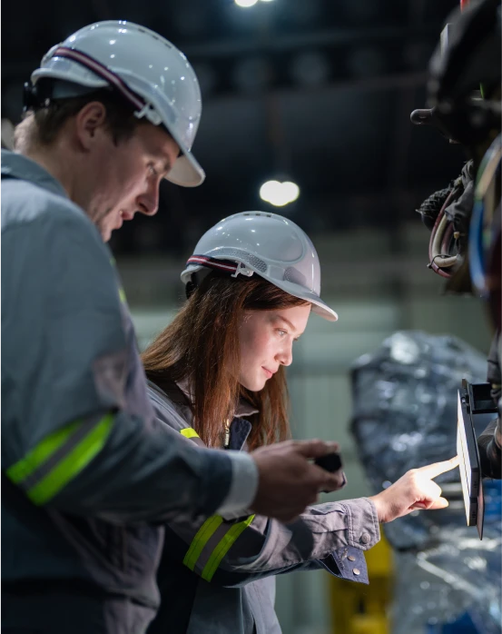 Deux travailleurs en tenue de sécurité et casque blanc inspectant un écran dans une usine.